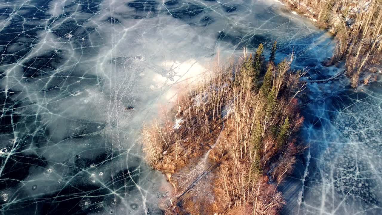 マタヌスカ湖上空を降下する空撮ドローン映像。ごつごつした枯れ木と風化した枝が点在する小さな岩だらけの島が現れる。その下には凍てついた水面が果てしなく広がり、深い青色をしている。