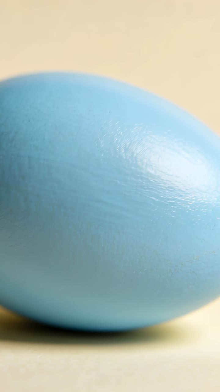 Vertical video: Pulling back camera, blue egg moving on pale table, showing profile over beige wall