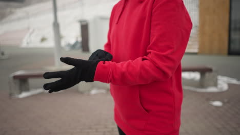 woman putting on black winter glove for protection against cold outdoors, background shows modern benches and light pole, emphasizing winter urban landscape, she is dressed in a cozy red hoodie