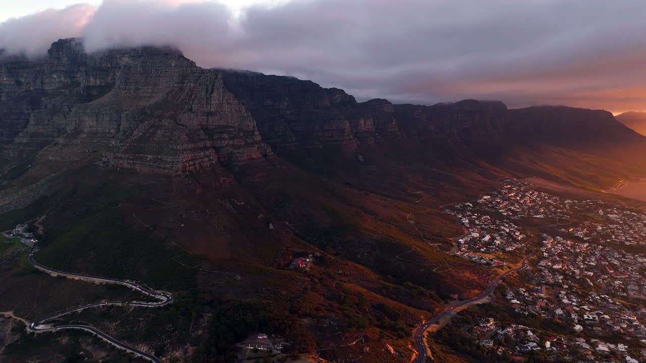Veduta aerea cinematografica della Table Mountain a Città del Capo, Sudafrica, scogliere rocciose drammatiche con paesaggio urbano costiero al tramonto, nuvole che si muovono sulle cime e luce dorata della sera.