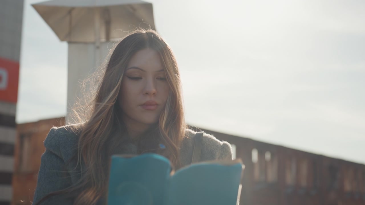 Bella donna che legge all'aperto sotto una luminosa luce solare in controluce. Scena urbana su un tetto con una giovane donna in cappotto di lana che sfoglia le pagine di un diario blu, con un morbido bagliore dorato, lens flare ed espressione contemplativa.