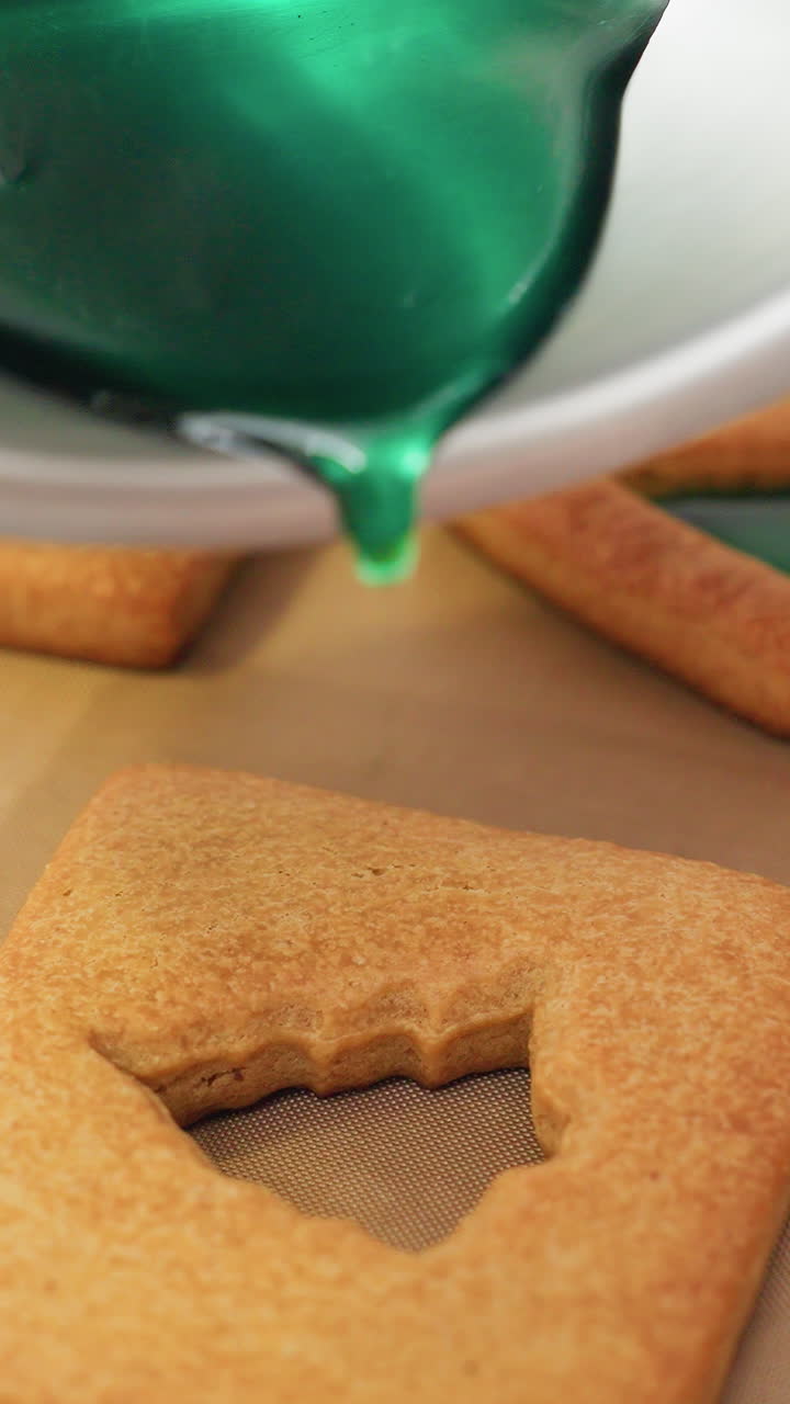 Close-up of vibrant green syrup being poured into the hollow center of shaped cookies, creating a decorative stained-glass effect on a parchment-lined baking tray in a professional kitchen setting