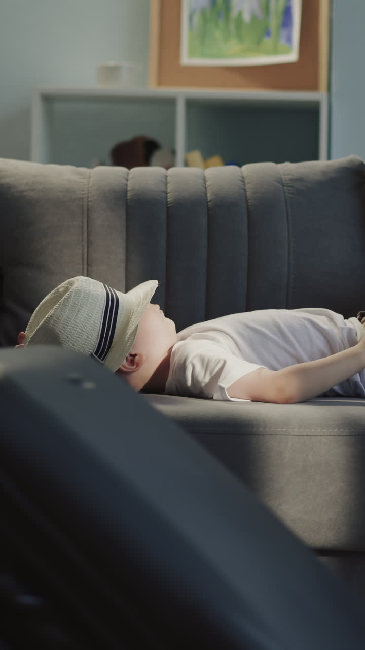 Little girl with braided hair pulls suitcase around sofa with lying toddler brother in living room. Funny naughty children ready for summer travel