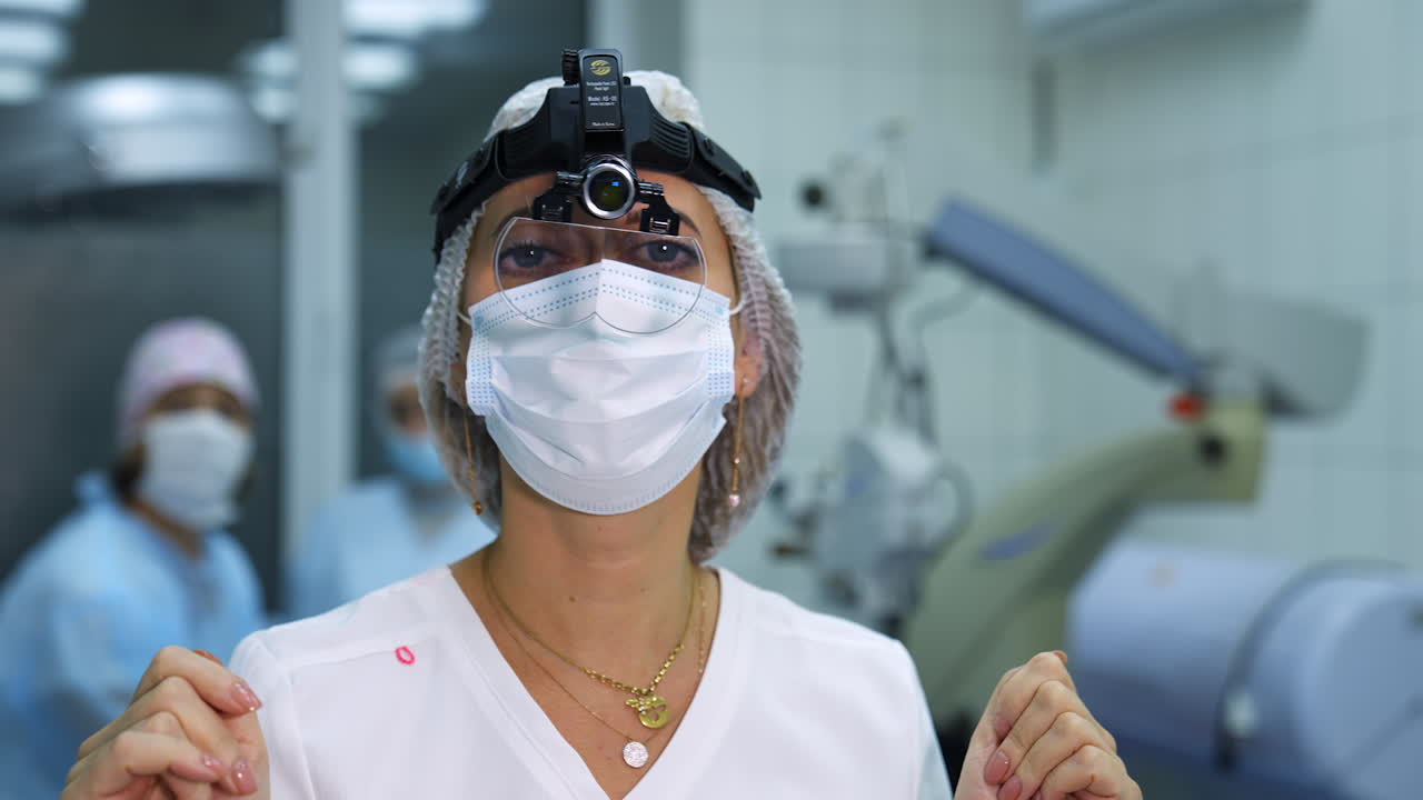 Ophthalmologist in magnifying glass on head in hospital. Portrait of a female doctor in a medical mask with professional lenses equipment in operating room.
