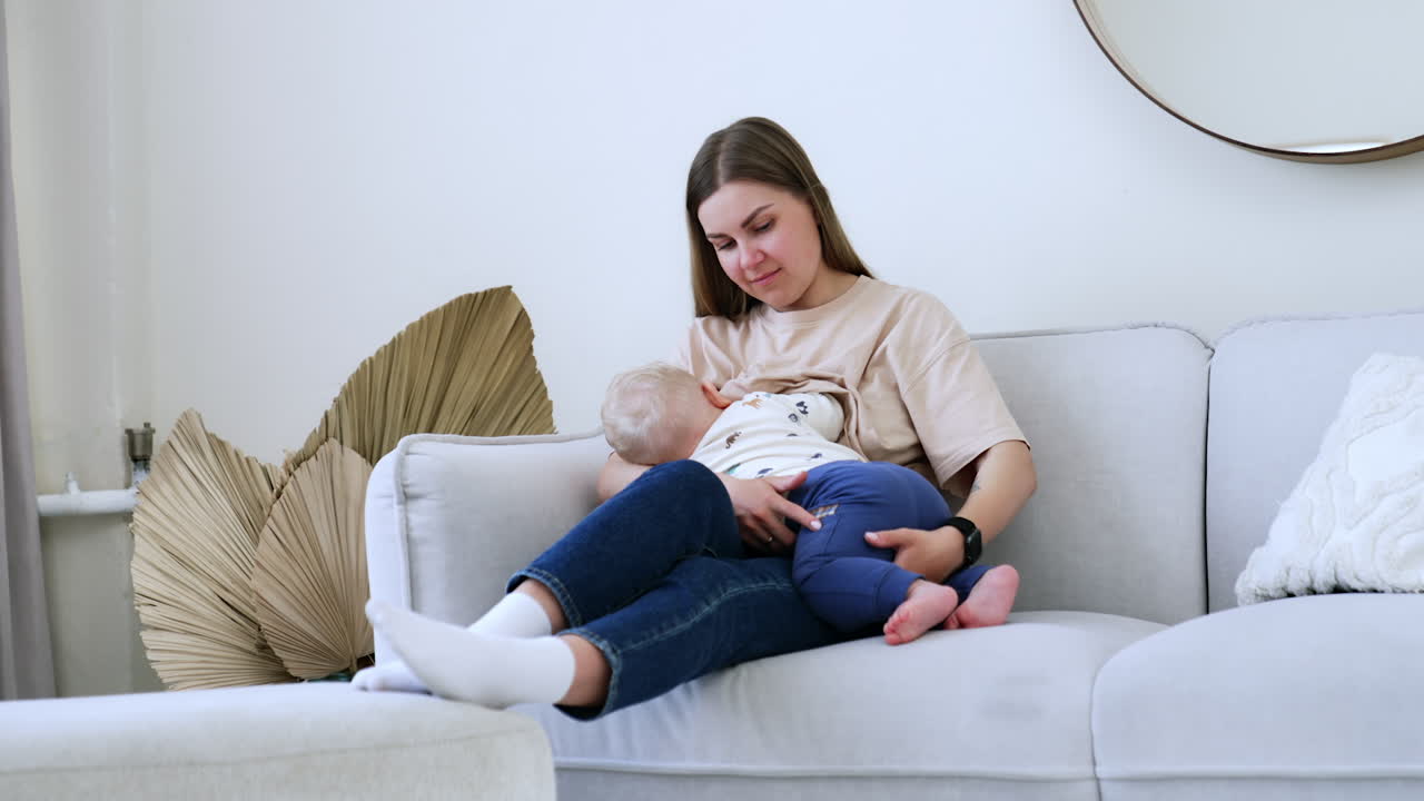 Long-haired Caucasian woman sits on the sofa. Mother is breastfeeding her infant baby boy.