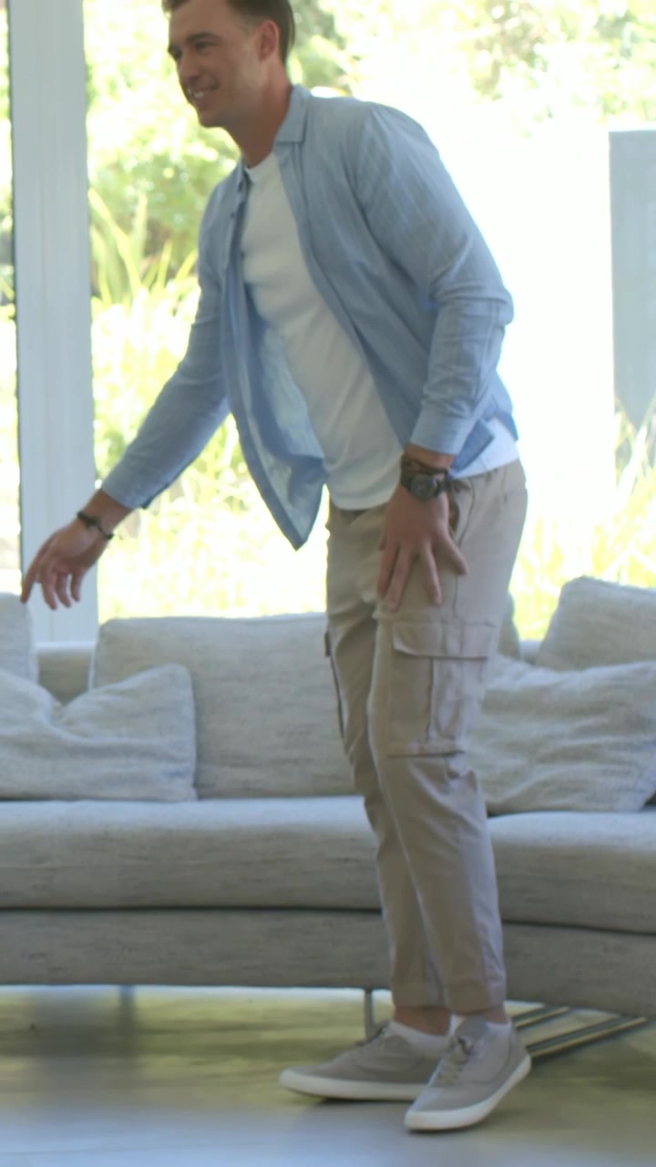 Vertical video: Entering room, man blue-shirt walking to sofa, reaching pillow to sit by glass-wall