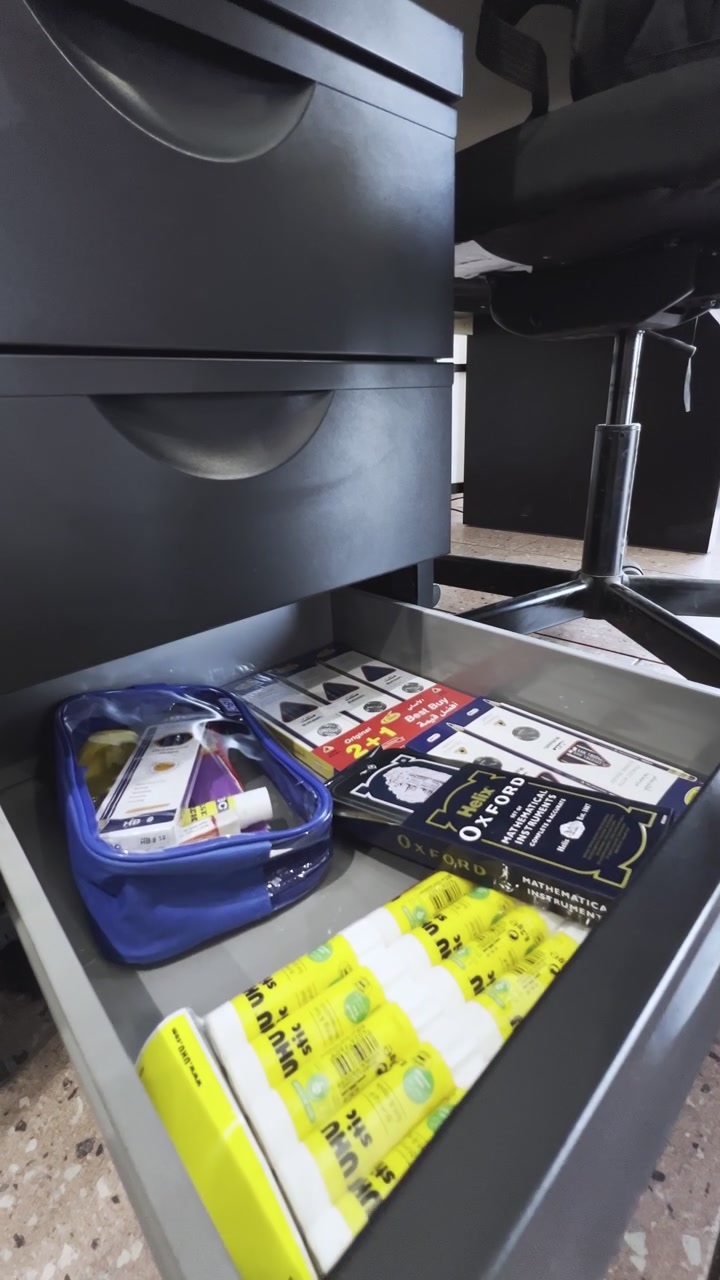 A close-up view of a hand placing a blue pencil case into an open desk drawer. The indoor office setting includes a black filing cabinet, chair wheels, and new school stationery supplies.