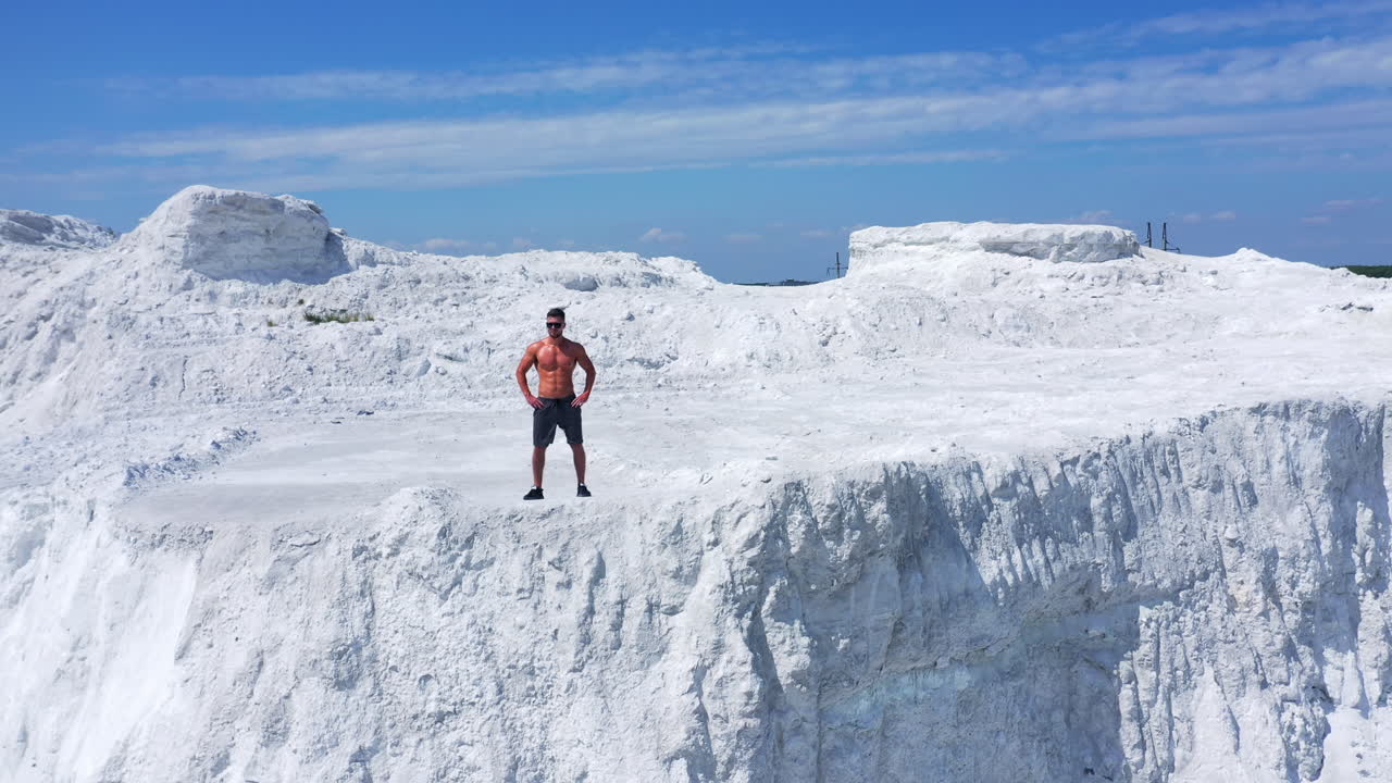 Drone view of a strong man in shorts without shirt on white hill. Young muscular man in good physical shape wearing sunglasses standing in nature. Camera moves up.