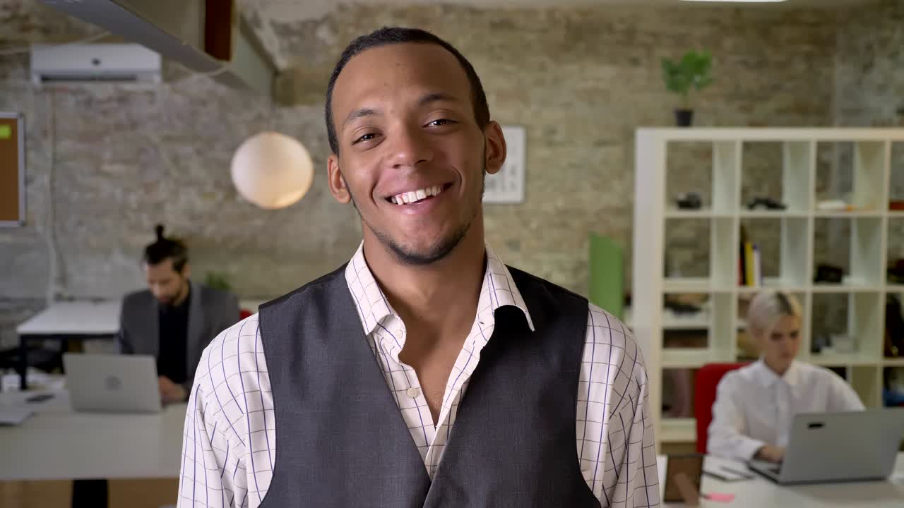 jovem empresário africano está de pé e olhando para a câmera no escritório, rindo, colegas estão trabalhando com laptops, conceito de trabalho