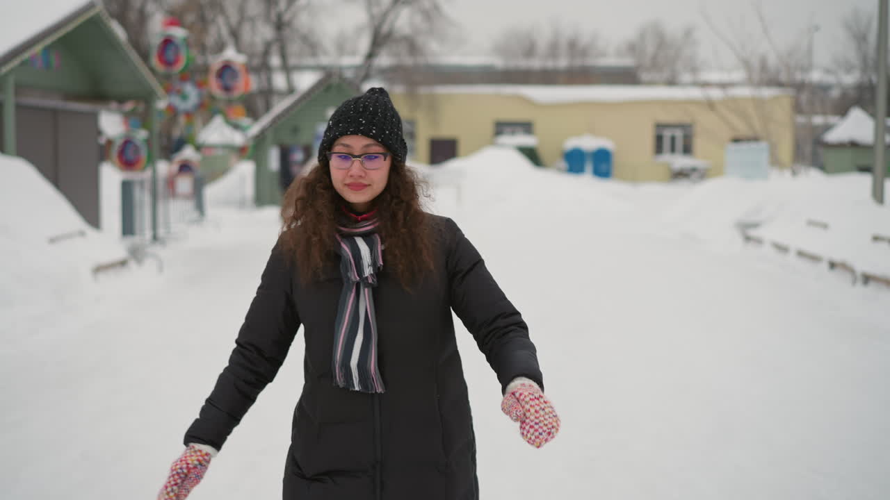 Athletic girl wearing winter jacket, knitted hat, scarf, and colorful mittens outdoors on snowy street in cold season, posing confidently with hand near face, enjoying fresh air and frosty atmosphere