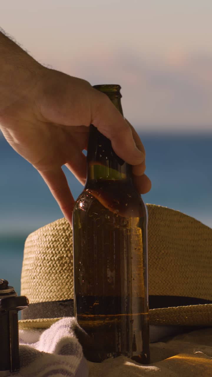 video concepto de vacaciones de verano vertical de botella de cerveza en la toalla de playa con sombrero de sol contra el cielo al atardecer 1