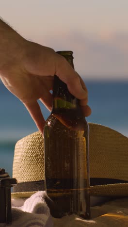 Vertical-Video-Summer-Holiday-Concept-Of-Beer-Bottle-On-Beach-Towel-With-Sun-Hat-Against-Sunset-Sky-1