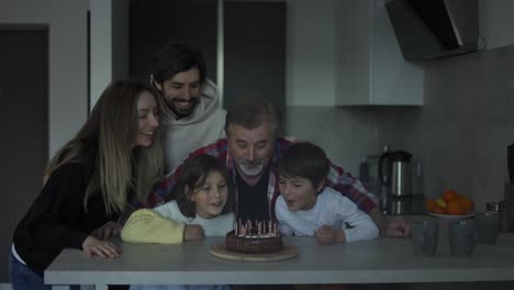 Abuelo-Feliz-Soplando-Velas-En-El-Pastel-De-Cumpleaños-Rodeado-De-Su-Familia