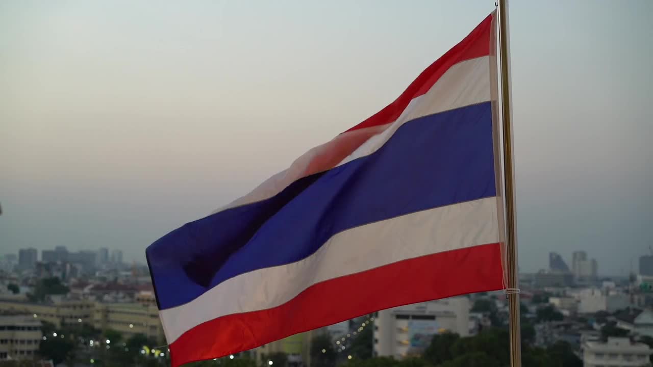 Thai flag waves atop rooftop with blurred urban skyline backdrop in Bangkok Thailand, symbolizing national pride and identity during evening with soft dusky light