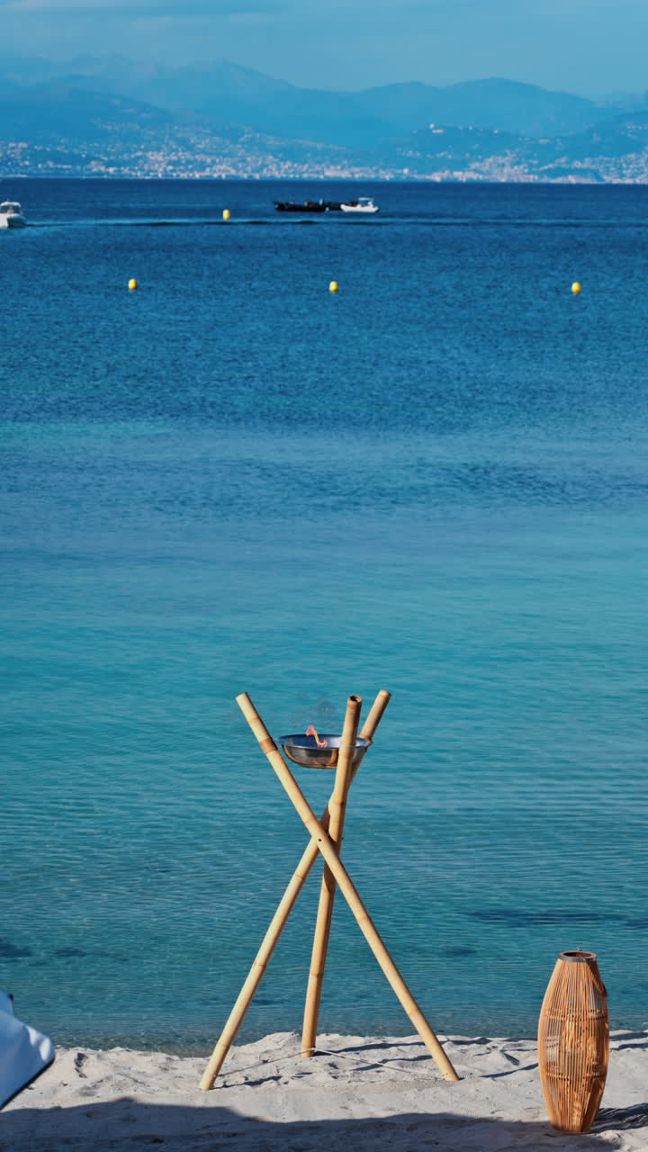 Bamboo structure holding a metal bowl with a small flame burning on the beach with the blue sea on the background. Vertical