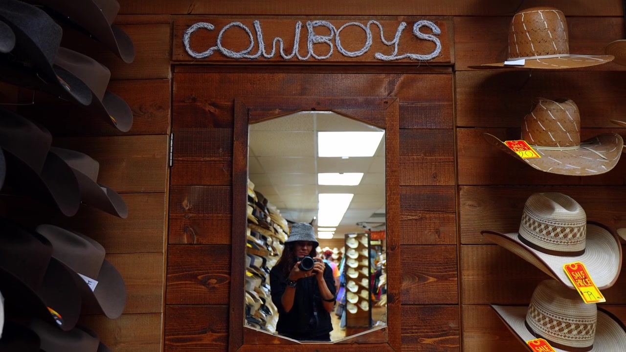 Esposizione di cappelli da cowboy in negozio con specchio e insegna con scritte in corda. Cappelli di paglia e feltro montati su parete di legno con cartellini del prezzo e luce interna calda.