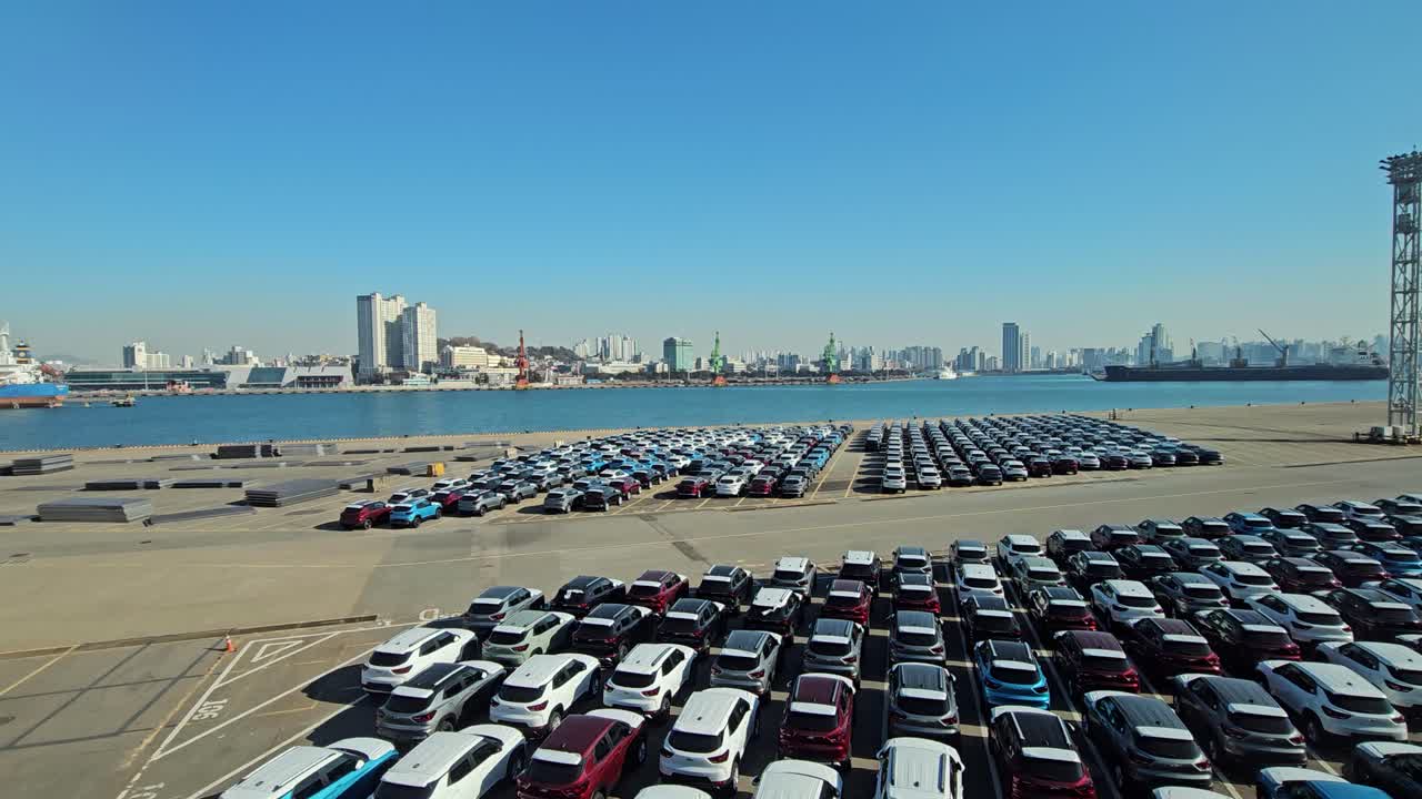 Rows of brand-new export cars are parked at Wolmi Port, Incheon, with the city skyline and docked ships creating a striking backdrop under a clear sky - dolly aerial