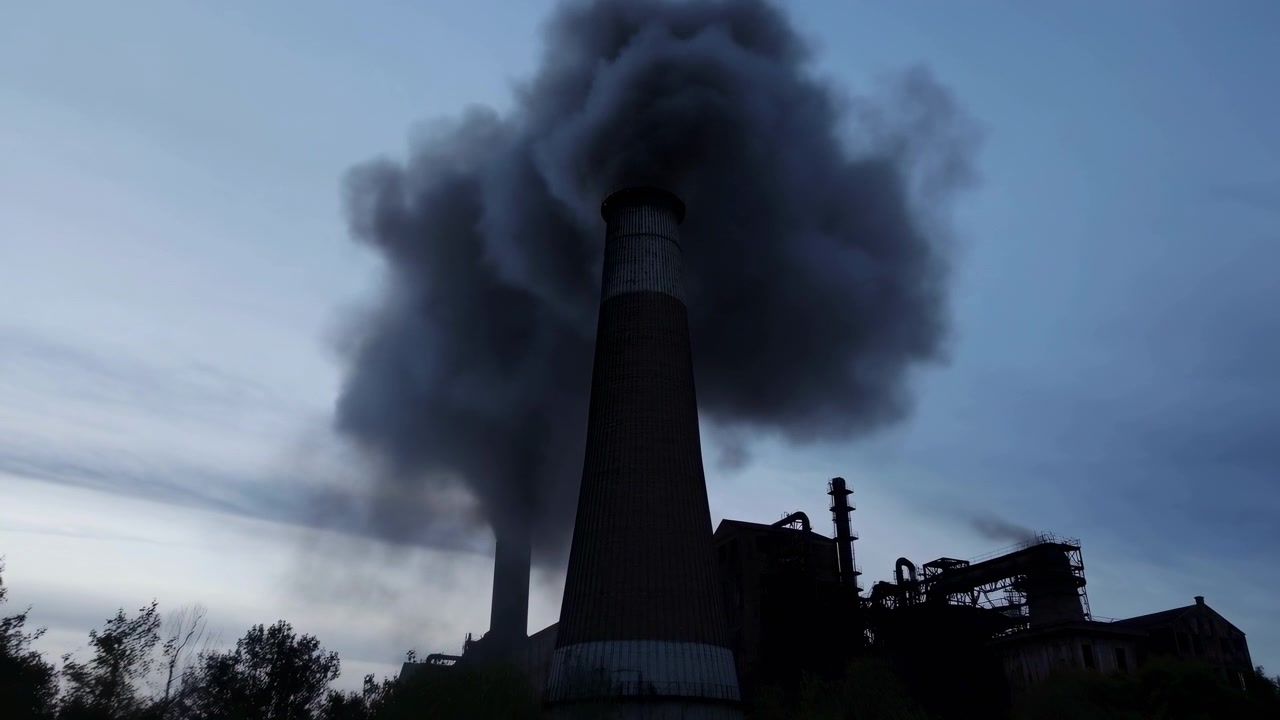 Scena industriale a livello del suolo che mostra un'alta ciminiera che rilascia fumo nero denso che sale e si diffonde nel cielo crepuscolare blu-grigio sopra edifici di fabbriche, tubi e strutture in silhouette