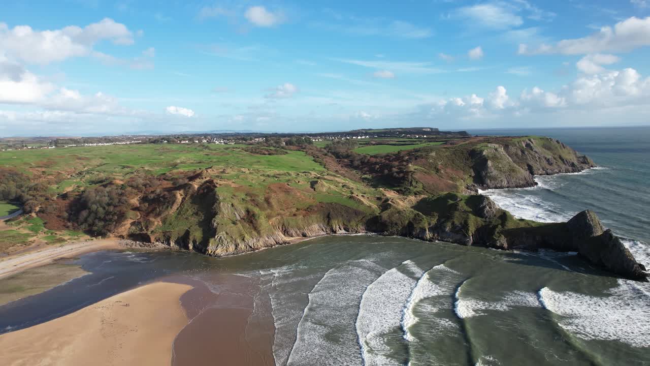 Aerial view of Three Cliffs Bay showing sweeping green cliffs rocky beach and rolling waves under clear summer skies on the Wales coastline
