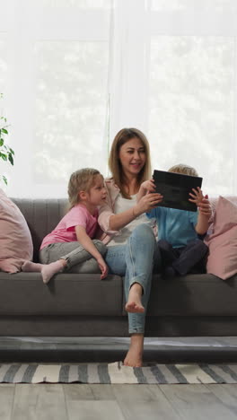 mother shows curious children interesting cartoons on tablet sitting on couch against bright window. toddler boy tries to take gadget to hold