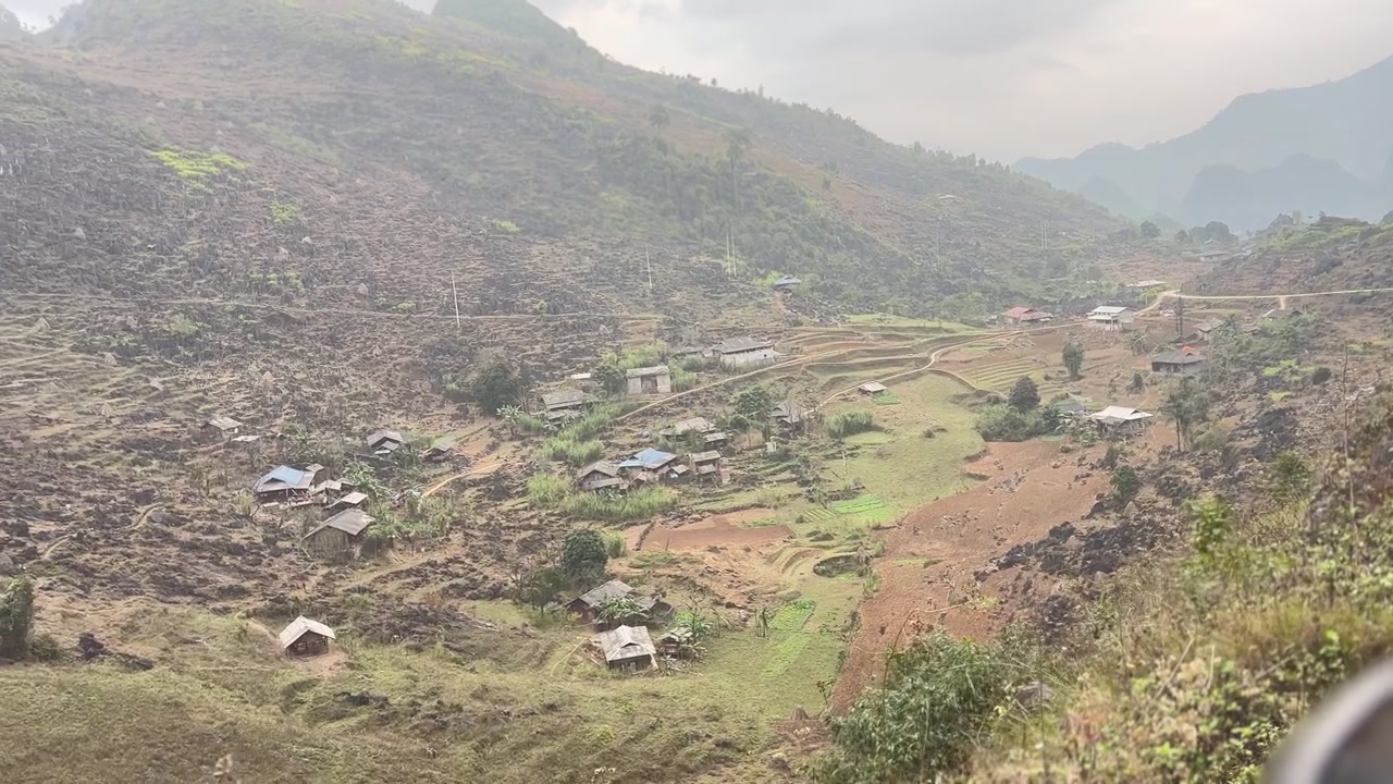 Vista dall'alto da una strada di montagna che domina un piccolo villaggio, campi terrazzati e un paesaggio di valle accidentato sotto cime nebbiose ad Ha Giang, Vietnam