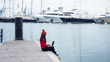 Asian-woman-sits-by-the-water-at-the-port-of-Valencia-ES,-engrossed-in-her-work-on-a-laptop,-her-thoughts-consumed-by-the-task-at-hand,-wearing-a-red-beanie-that-complements-her-red-turtleneck-shirt