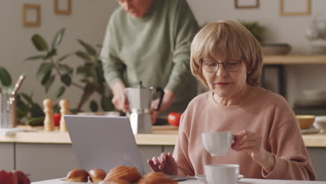 senior woman using laptop, drinking coffee and talking with husband