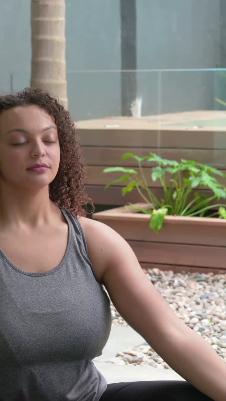 Vídeo vertical: Mulher de regata cinza olhando para cima, elevando as palmas das mãos em gesto de acalmar-se, no pátio, com plantas.