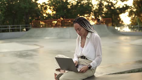 Young-elegant-caucasian-woman-with-dreadlocks-in-white-blouse-sitting-in-summer-park.-Happy-freelancer-typing-on-laptop-outdoors