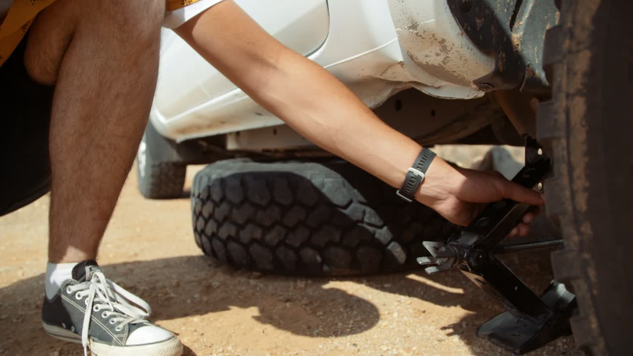 Dynamic tracking low wide hand held shot of a Caucasian male tourist in Africa as he removes the car jack from the underneath of a off-roading vehicle after having changed the tire