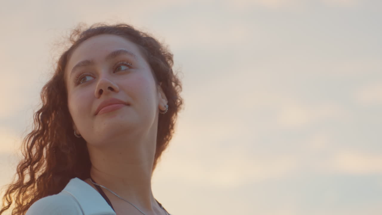 Retrato de mujer juguetona bajo un cielo dorado en una isla, miradas sonrientes y movimiento espontáneo, brisa costera a través del cabello rizado, piel bronceada por el sol y pose relajada, ambiente de viaje de estilo de vida con el resplandor del atardecer.