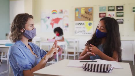 maestra y niña con máscaras faciales hablando entre sí a través del lenguaje de señas en la clase