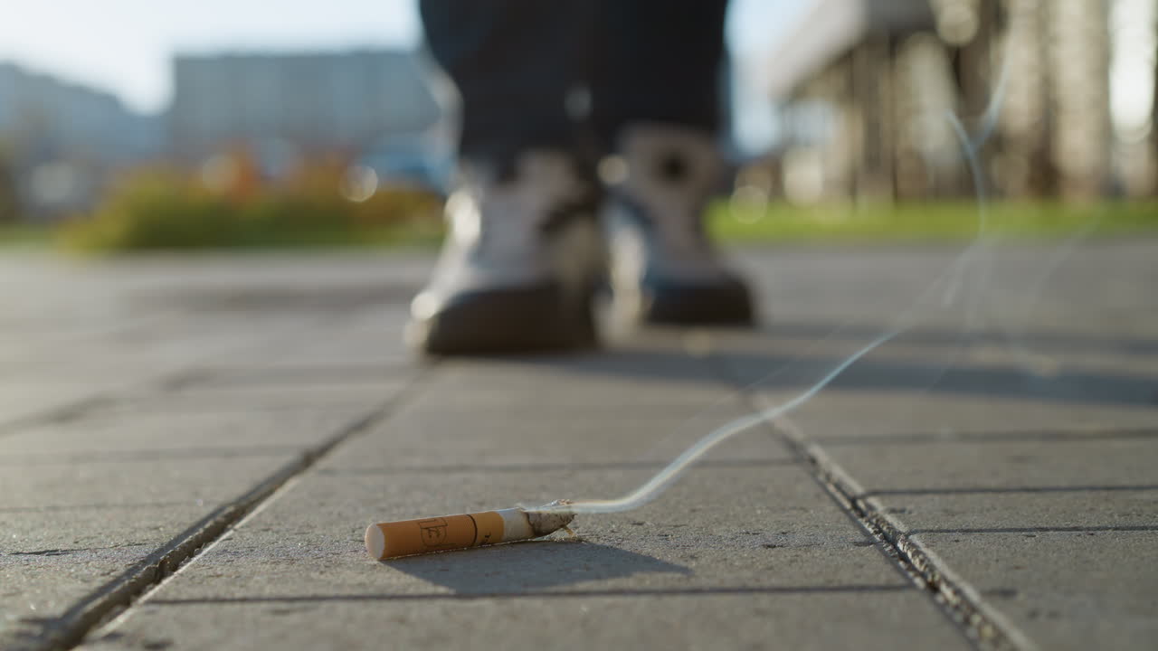 close up of burning cigarette lying on pavement with swirling smoke rising as blurred figure in background approaches slowly, suggesting intent or reaction to discarded tobacco in urban setting