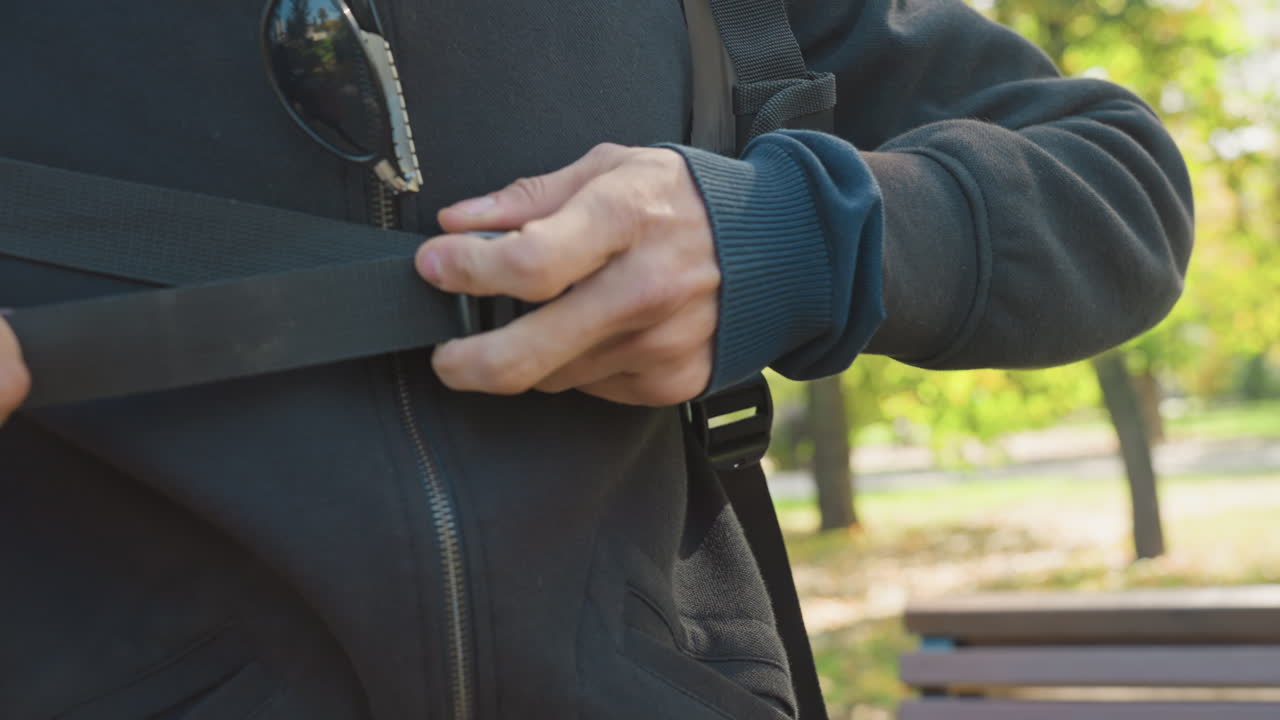Courier fastening shoulder strap in park hands adjusting buckle on black backpack strap over zippered jacket smartwatch visible bench and sunlit trees in background preparing for last mile delivery