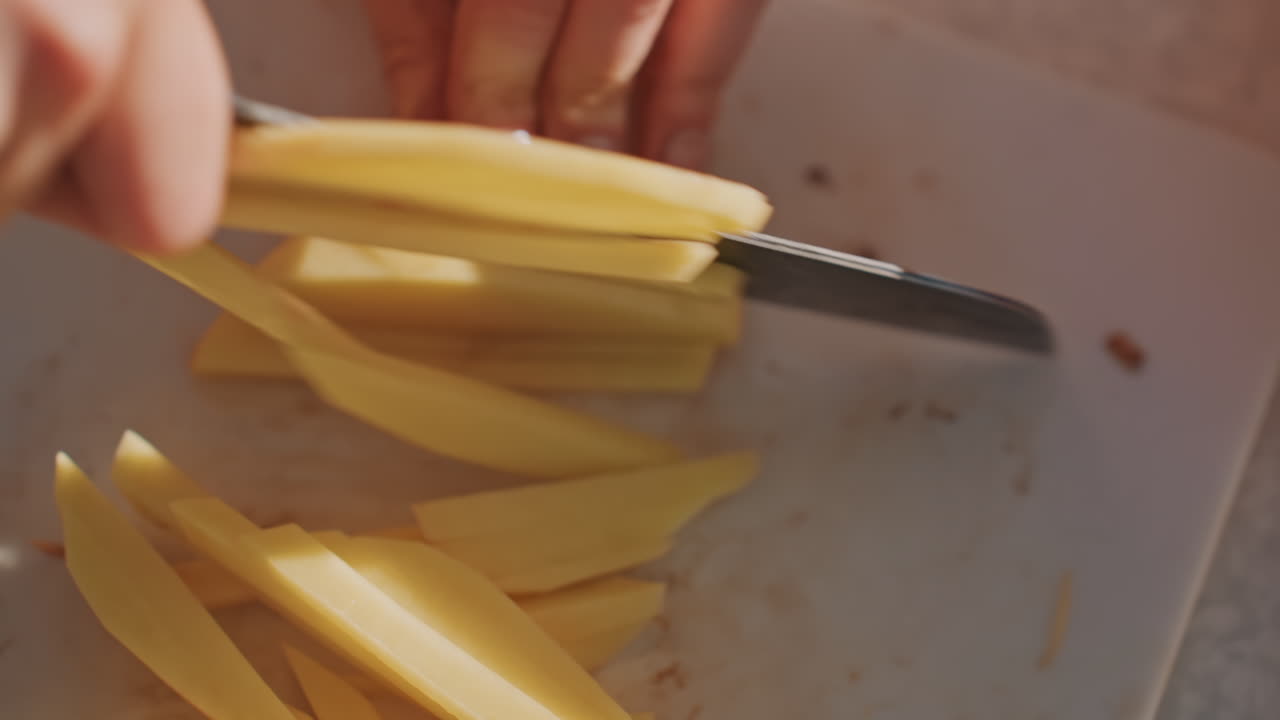 Focused hands cutting potato into fries on white board, even batons lined up in warm morning light, steady knife motion, campfire cookware nearby, meticulous backcountry meal prep