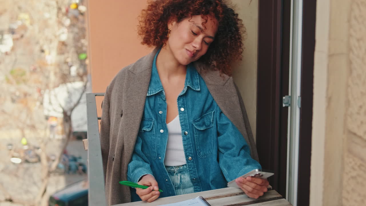 Young Woman Writing Notes and Using Smartphone on Balcony