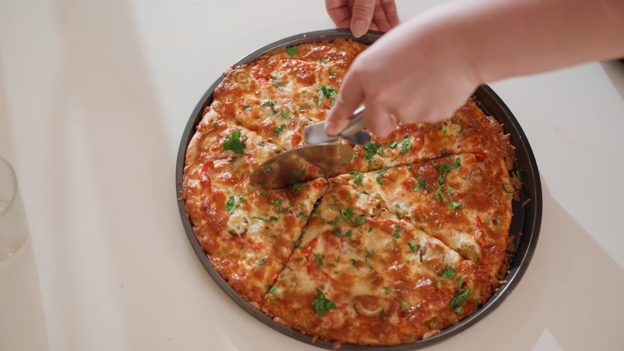 Vista dall'alto di una pizza rotonda cotta in una teglia di metallo, tagliata e servita da due mani; l'azione visibile mostra un tagliapizza e una spatola che affettano il formaggio fuso e la salsa di pomodoro mentre la spatola solleva una fetta.