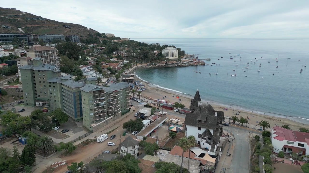 Volo in avanti sul centro di Papudo che rivela condomini, una cappella storica, una spiaggia sabbiosa, una baia calma con barche a vela ancorate e colline aride sotto un cielo coperto. Papudo, Valparaíso, Cile.