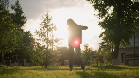 back view of lady joyfully dancing outdoors in grassy field with book in hand as sunlight creates radiant atmosphere around her, with a mix of greenery, trees, and building in background