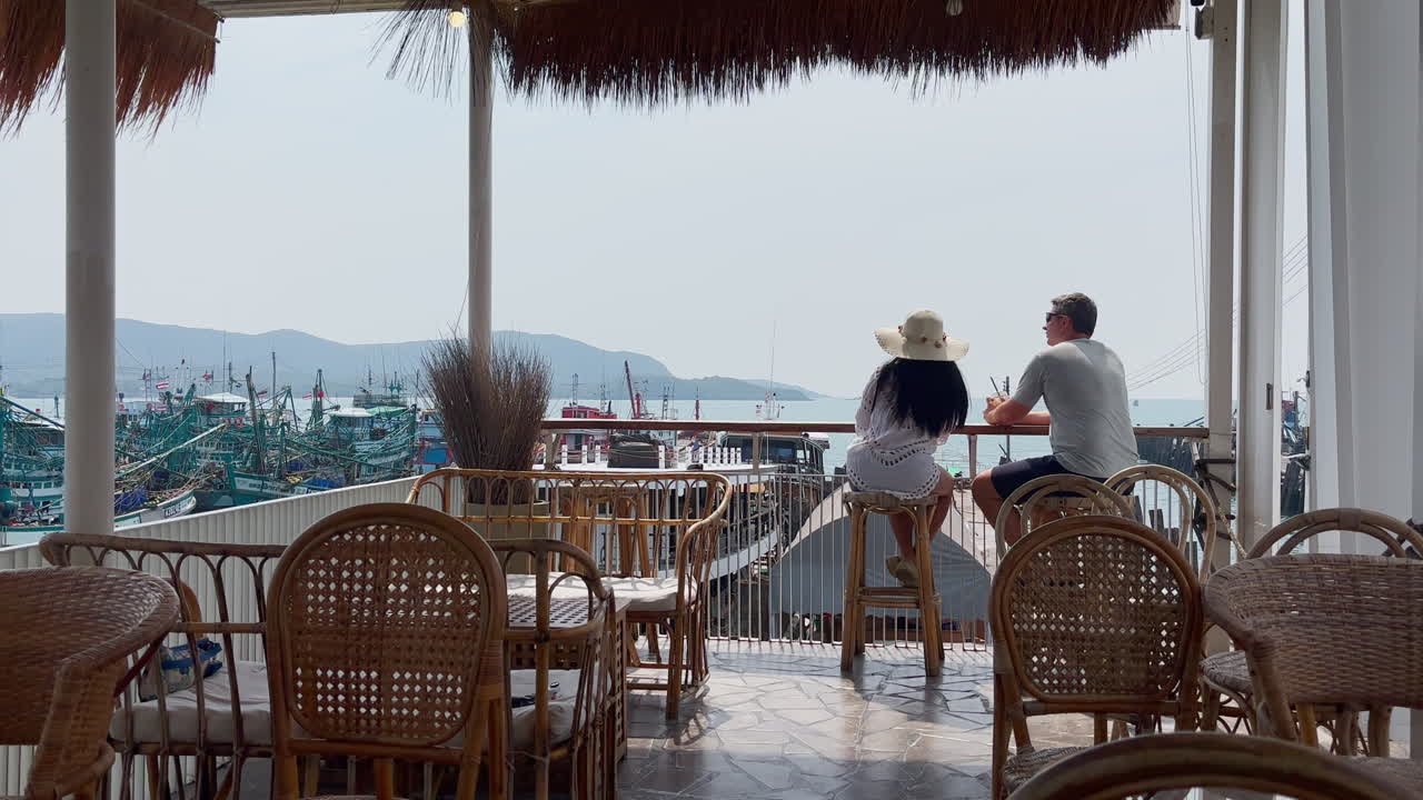 Paar entspannt in einem Hafen-Café in Thailand, genießt den Meerblick und die Küstenatmosphäre. Reiselifestyle-Szene mit Fischerbooten und friedlicher Küstenlandschaft in Südostasien.