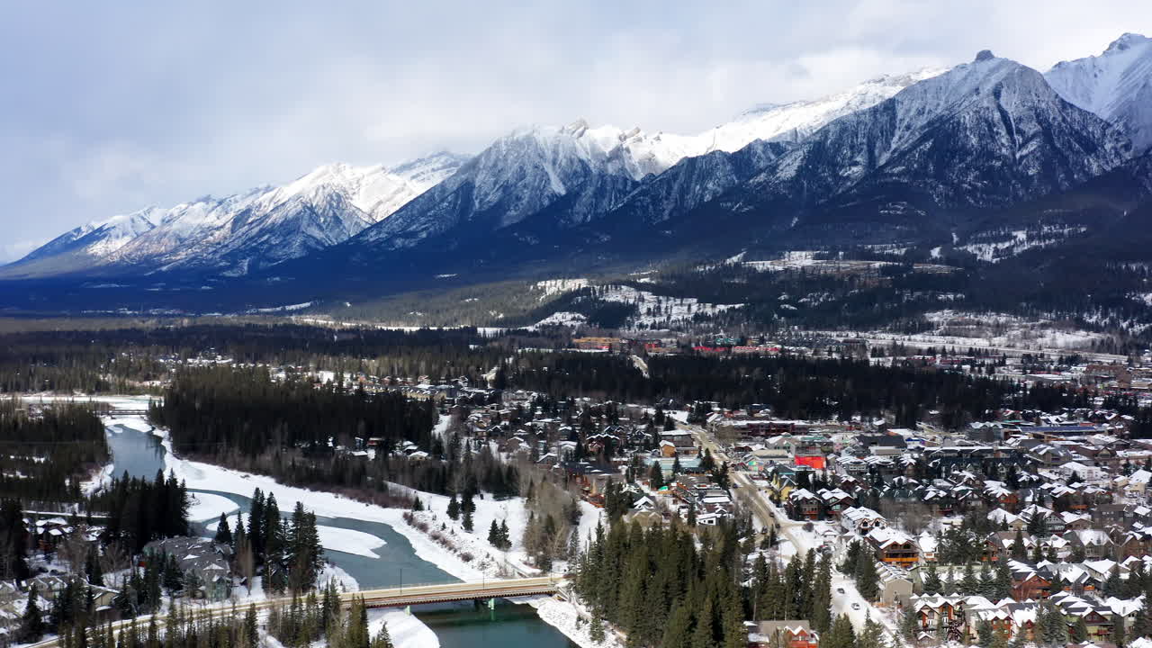 A drone pans and flies into the heart of snowy Canmore, revealing the town tucked beneath the towering peaks of the BANFF Mountains.