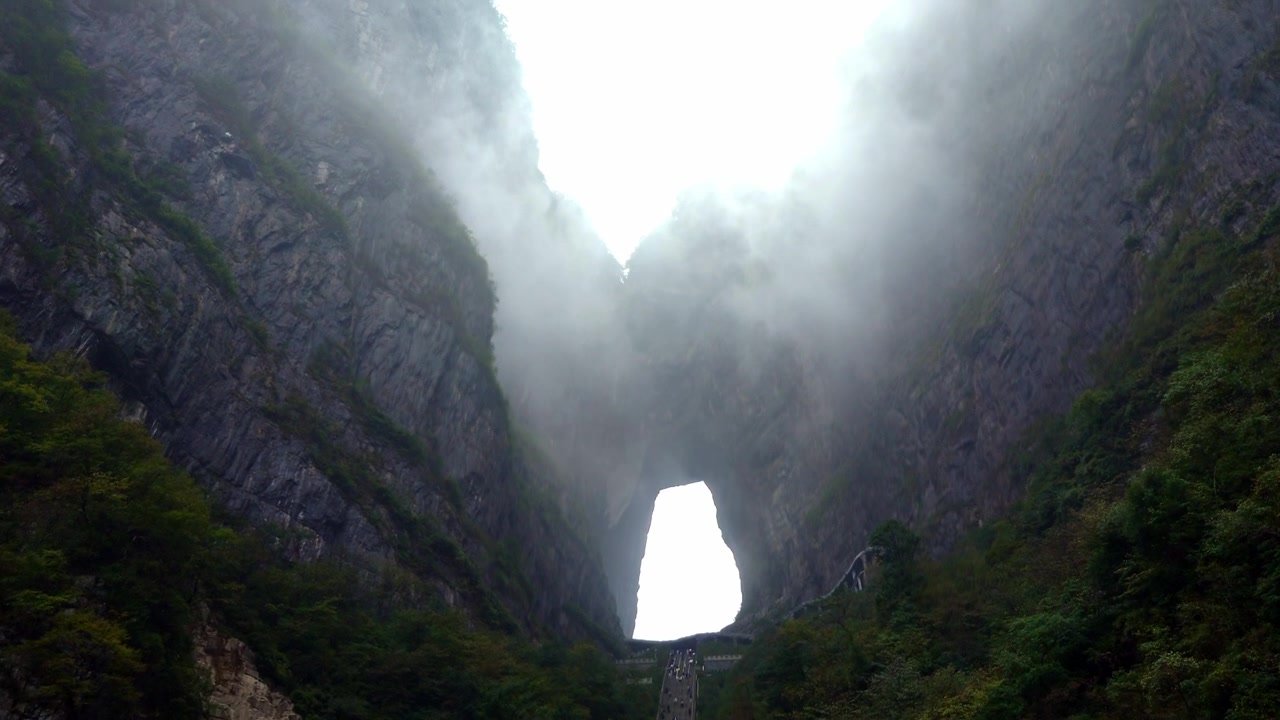 A dramatic low-angle shot of steep 999 steps staircase and the massive natural archway of Tianmen Cave on Tianmen Mountain. Thick, moody mountain fog rolls through the giant stone aperture.