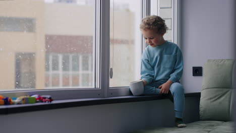 Un-Niño-Lindo-En-El-Alféizar-De-La-Ventana-En-La-Guardería-Un-Niño-Feliz-Con-El-Pelo-Rizado-Está-Bebiendo-Leche-Caliente-De-Una-Taza