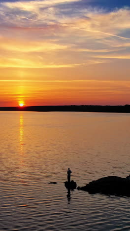 flying over the evening river and some people standing on a stone in water. golden sun setting on the natural water background. silhouette of a fisherman at sunset. vertical video