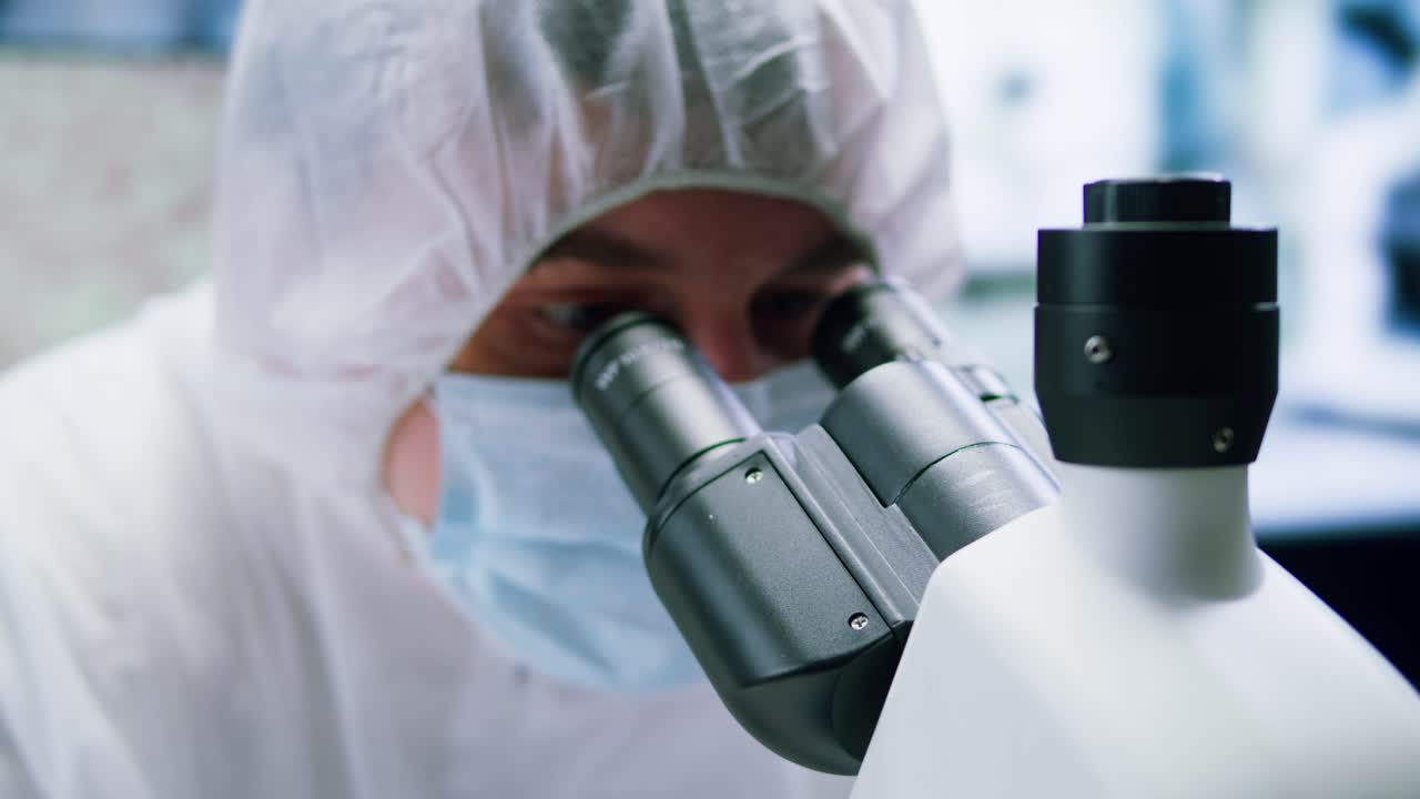 Scientist in Protective Gear Adjusting and Looking Through a Microscope