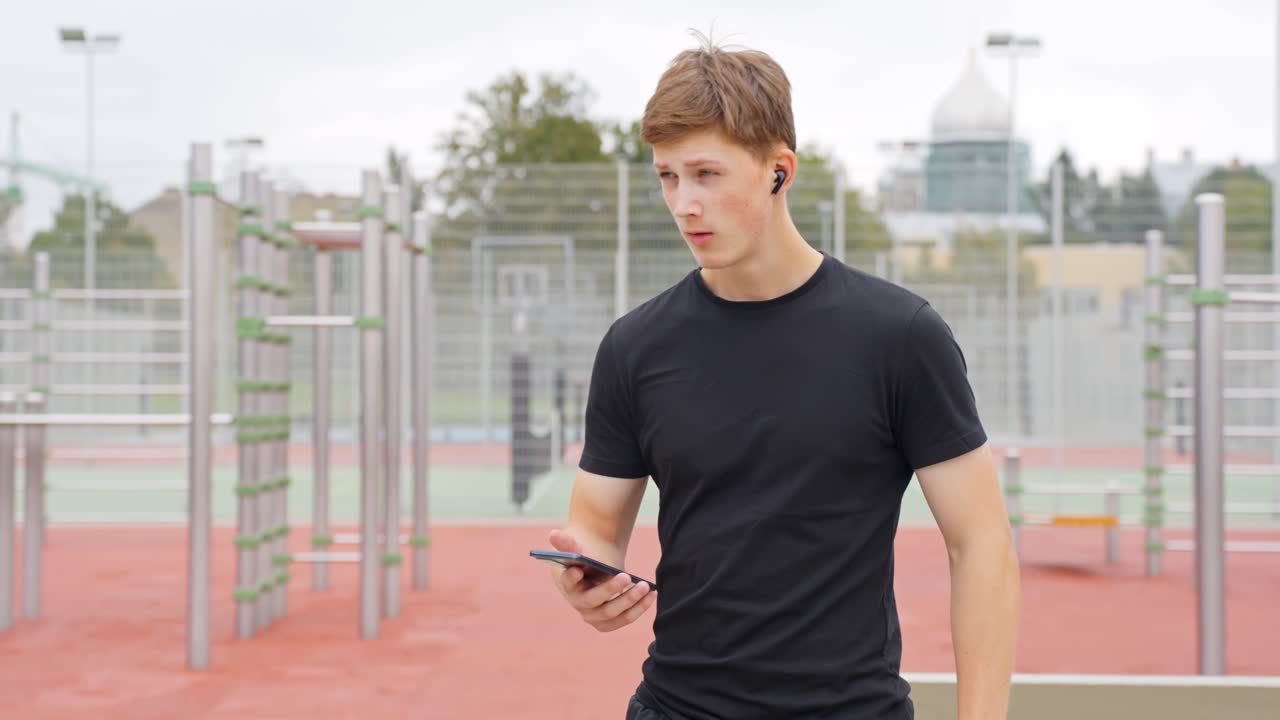 Man adjusts songs scrolling on phone while standing in sports area during workout break