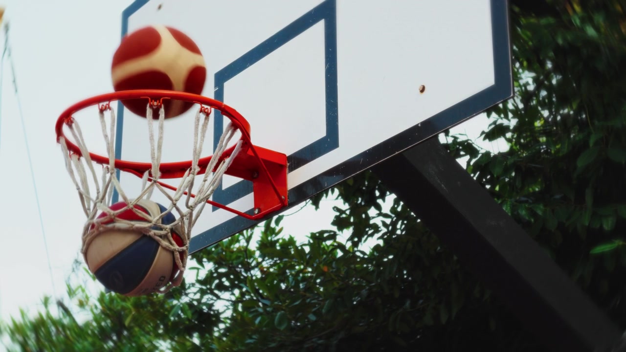 Palla da basket che cade attraverso un canestro esterno al rallentatore, circondata da alberi verdi
