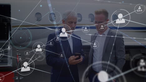 animation of network of profile icons over two caucasian businessmen using smartphone at airport