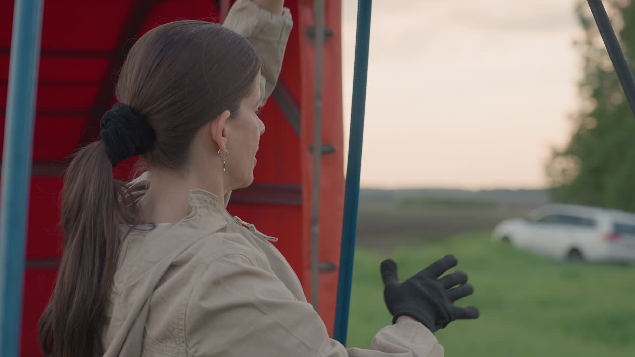 lady securing burner hoses and frame supports inside wicker basket of hot air balloon near parked car and red trailer in grassy field capturing preparation tasks for upcoming flight mission