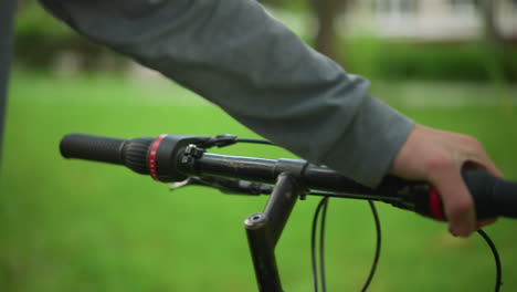 primer plano de una bicicleta estacionada con vista parcial de una mano con ropa gris sosteniendo el manillar de la bicicleta, observando la bicicleta, el fondo está borroso, con un exuberante parque verde